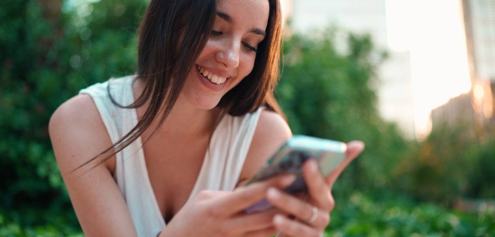 Woman outdoors smiling at her phone with greenery in the background.