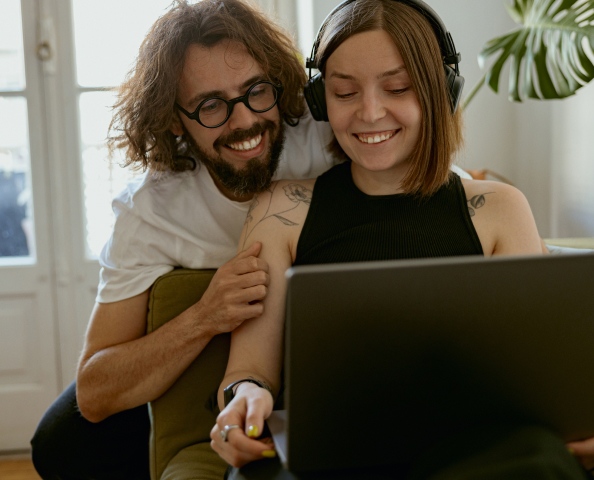 Two people smiling at a laptop screen; one is wearing headphones and the other is leaning on the couch.