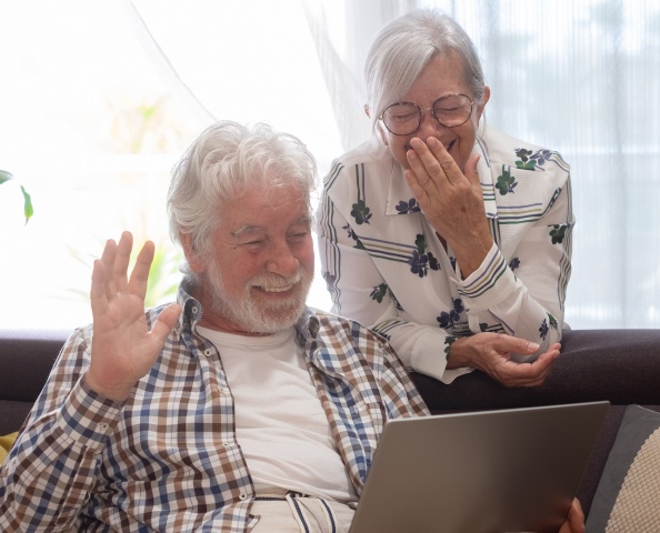 An elderly couple sits on a couch, smiling and using a laptop. The man waves while the woman covers her mouth with a hand, appearing amused.