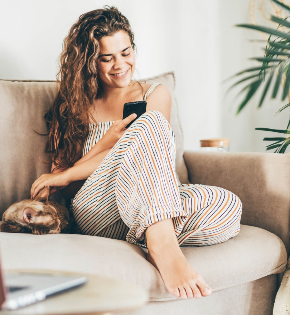 Woman sitting on a couch, smiling at her smartphone. She's wearing a striped outfit, and there's a cat lounging beside her. A plant is visible in the background.