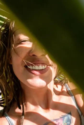 A person smiling behind a large leaf, partially obscured, with sunlight casting shadows on their face.