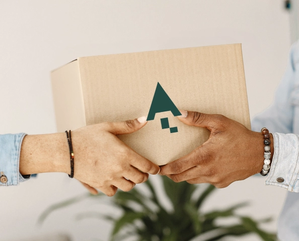 Two people holding a cardboard box with a green arrow logo. A plant is visible in the background.