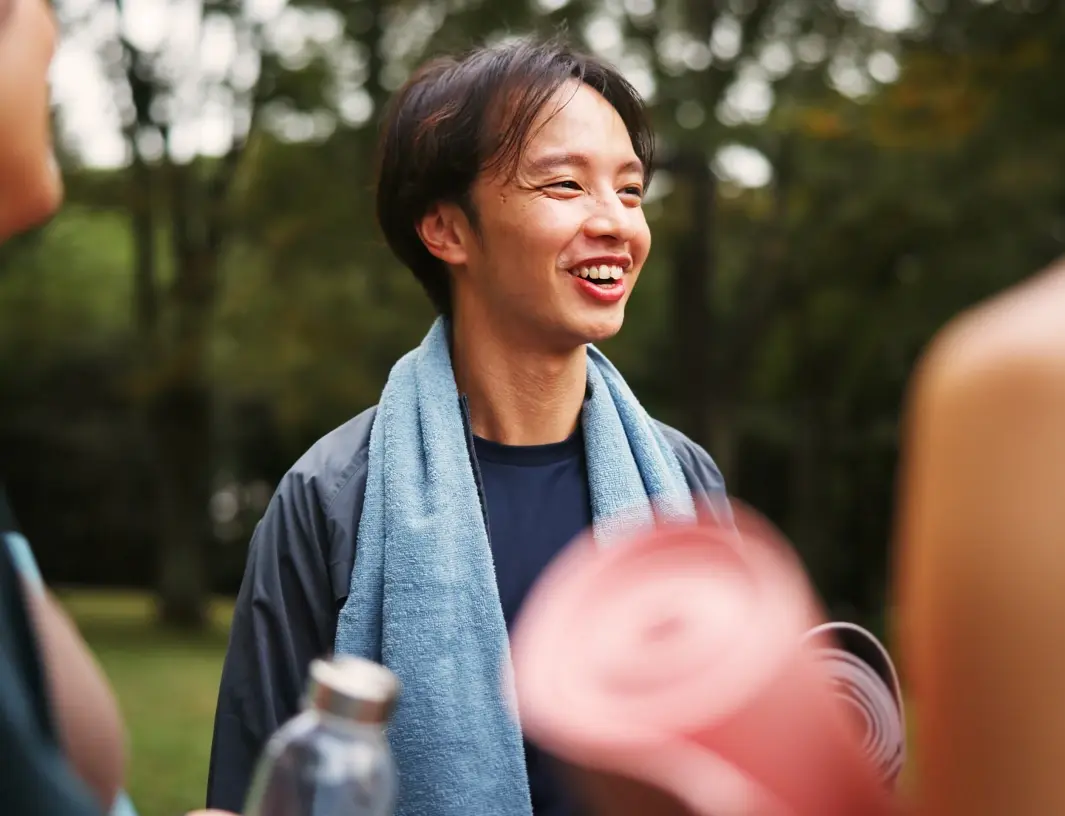 A person outdoors with a blue towel around their neck, smiling while holding a pink rolled-up mat.