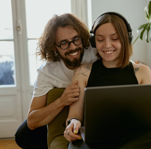 A person with long hair and glasses smiles while standing behind a seated person with headphones using a laptop.