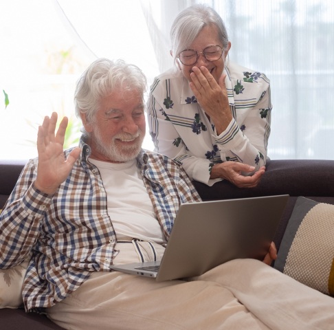 Elderly couple smiling and waving at a laptop screen while sitting on a sofa in a bright room.