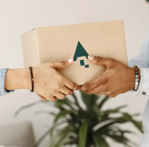 Two people exchange a cardboard box with a green triangle logo. A plant is blurred in the background.