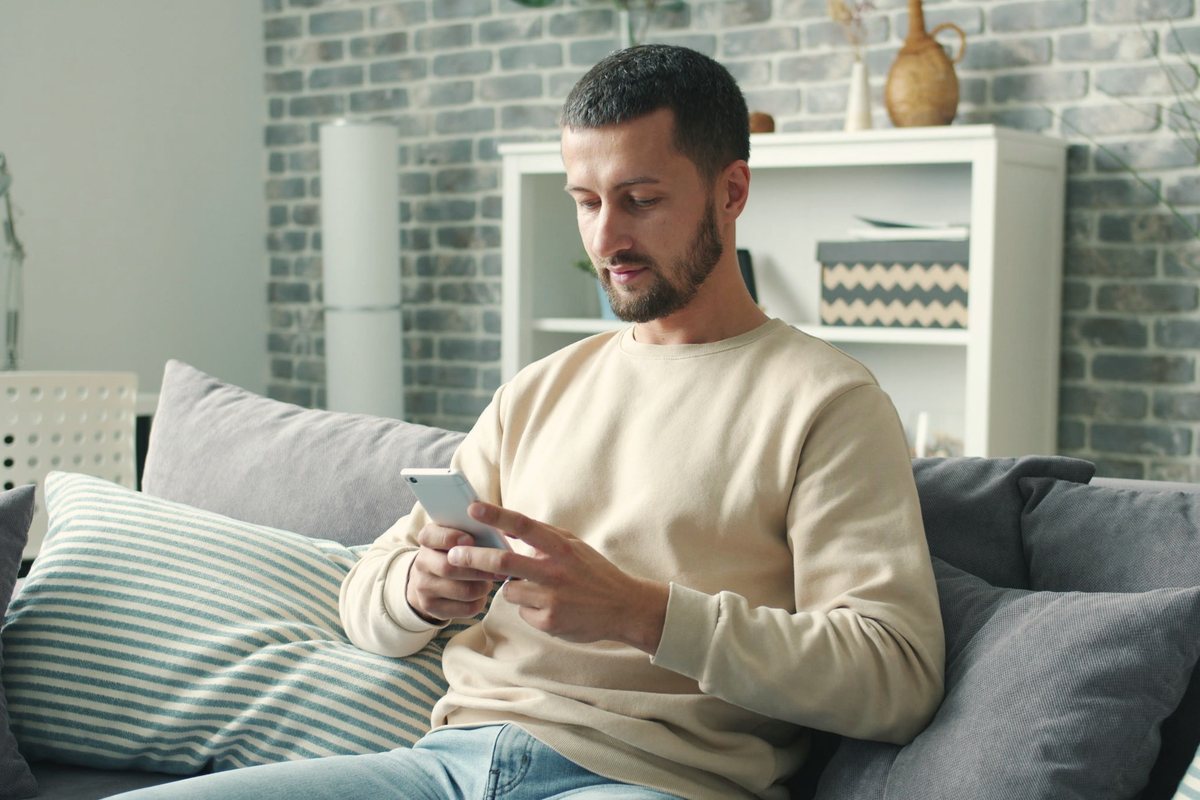 Man sitting on a grey couch at home using his phone to fill in an online booking form