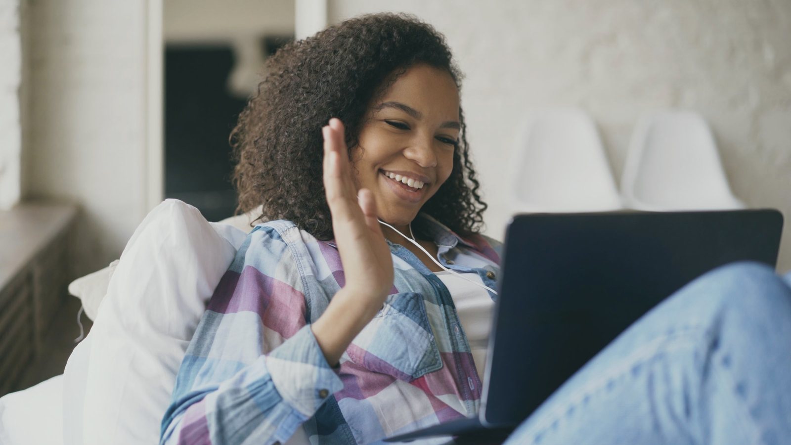 Smiling woman relaxing on a sofa, waving hello on a video call on her laptop