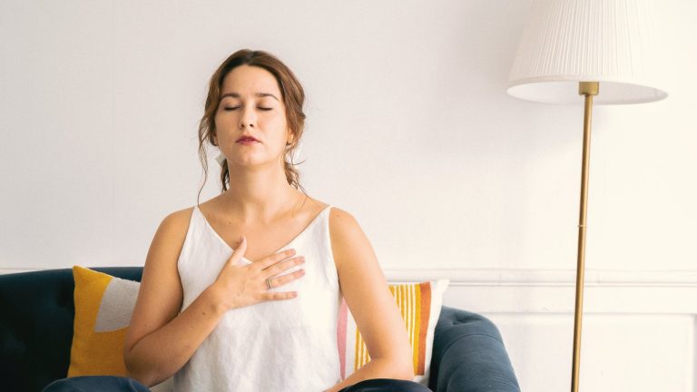 Woman sitting on a sofa with a hand on her chest and eyes closed, practising slow breathing