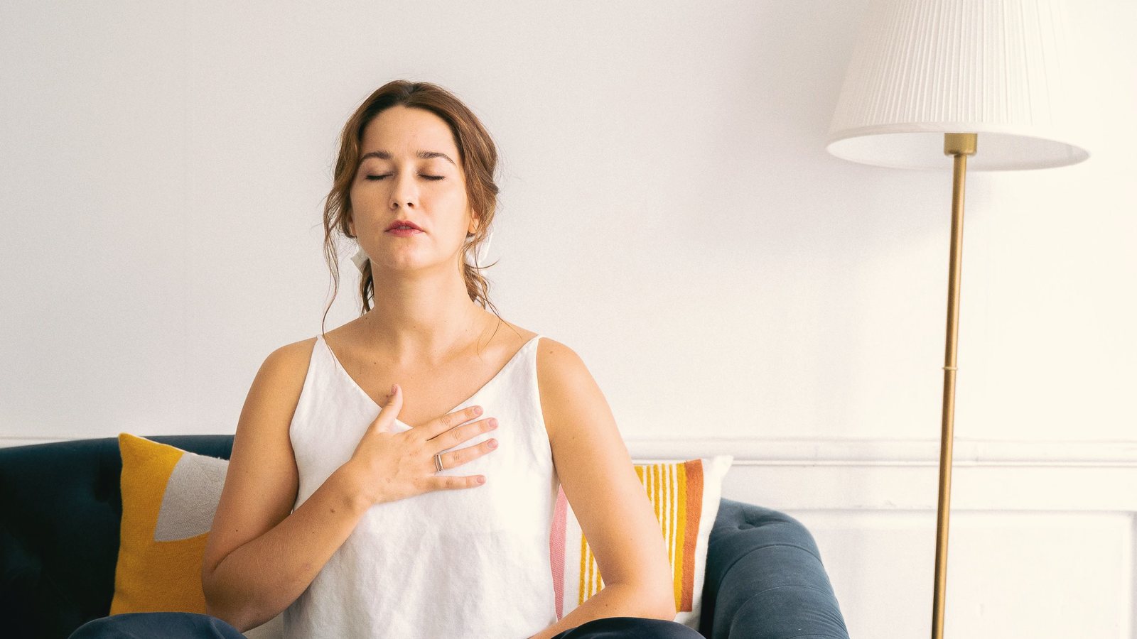 Woman sitting on a sofa with a hand on her chest and eyes closed, practising slow breathing