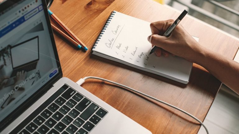 Person writing notes in a notepad beside an open laptop on a wooden desk