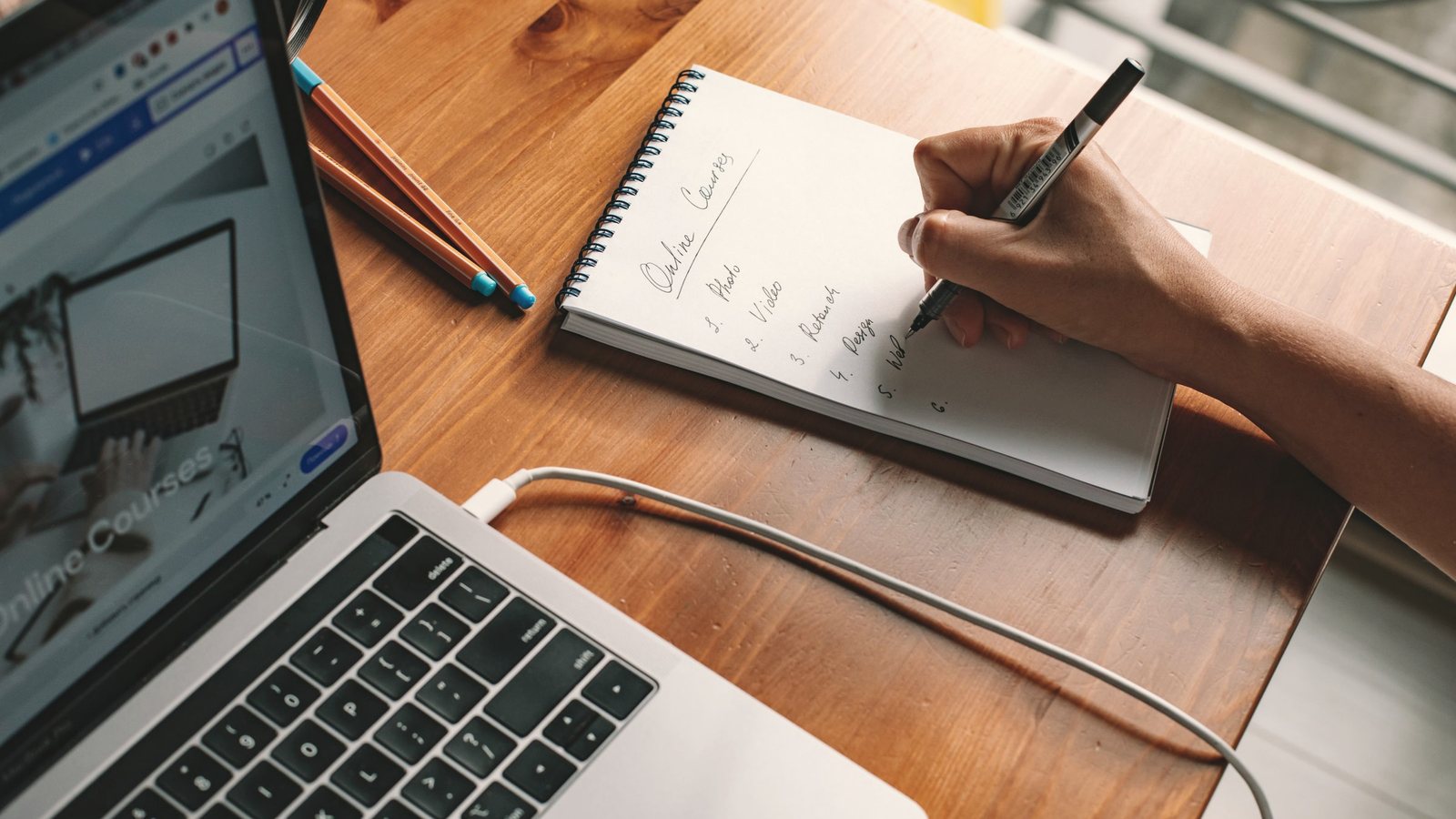 Person writing notes in a notepad beside an open laptop on a wooden desk