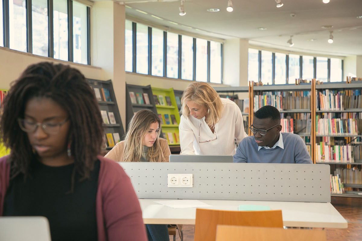Students working on a laptop in a library with a teacher