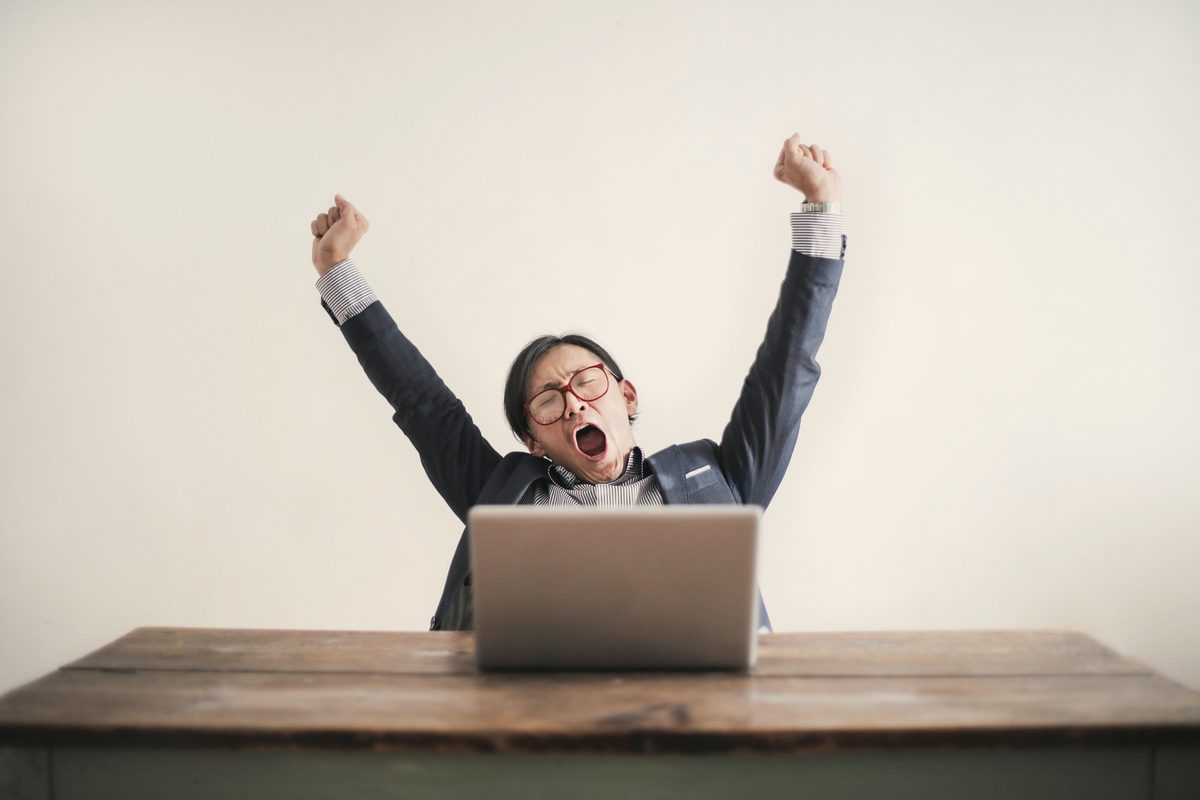 Yawning man with his hands up and eyes closed sitting at a table with his laptop