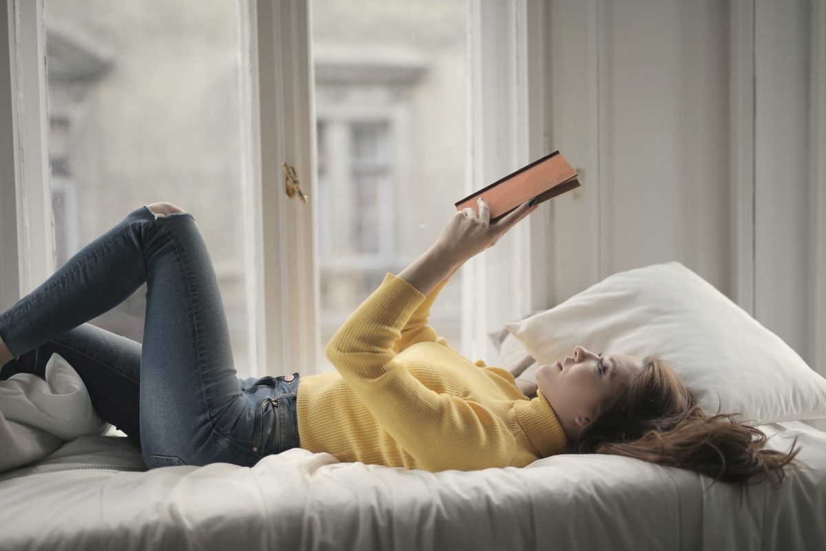 Woman in yellow sweater lying on bed while reading book