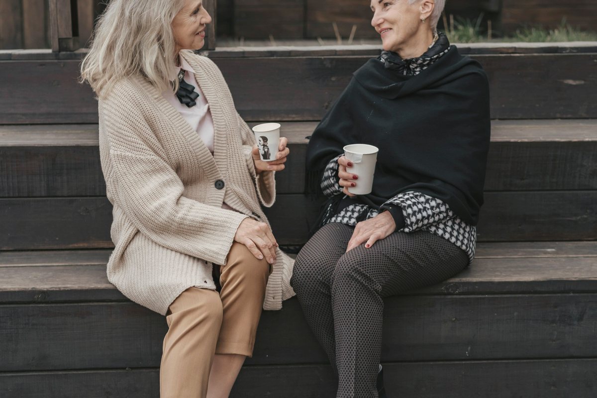 Two older women sitting on outdoor steps chatting over coffee