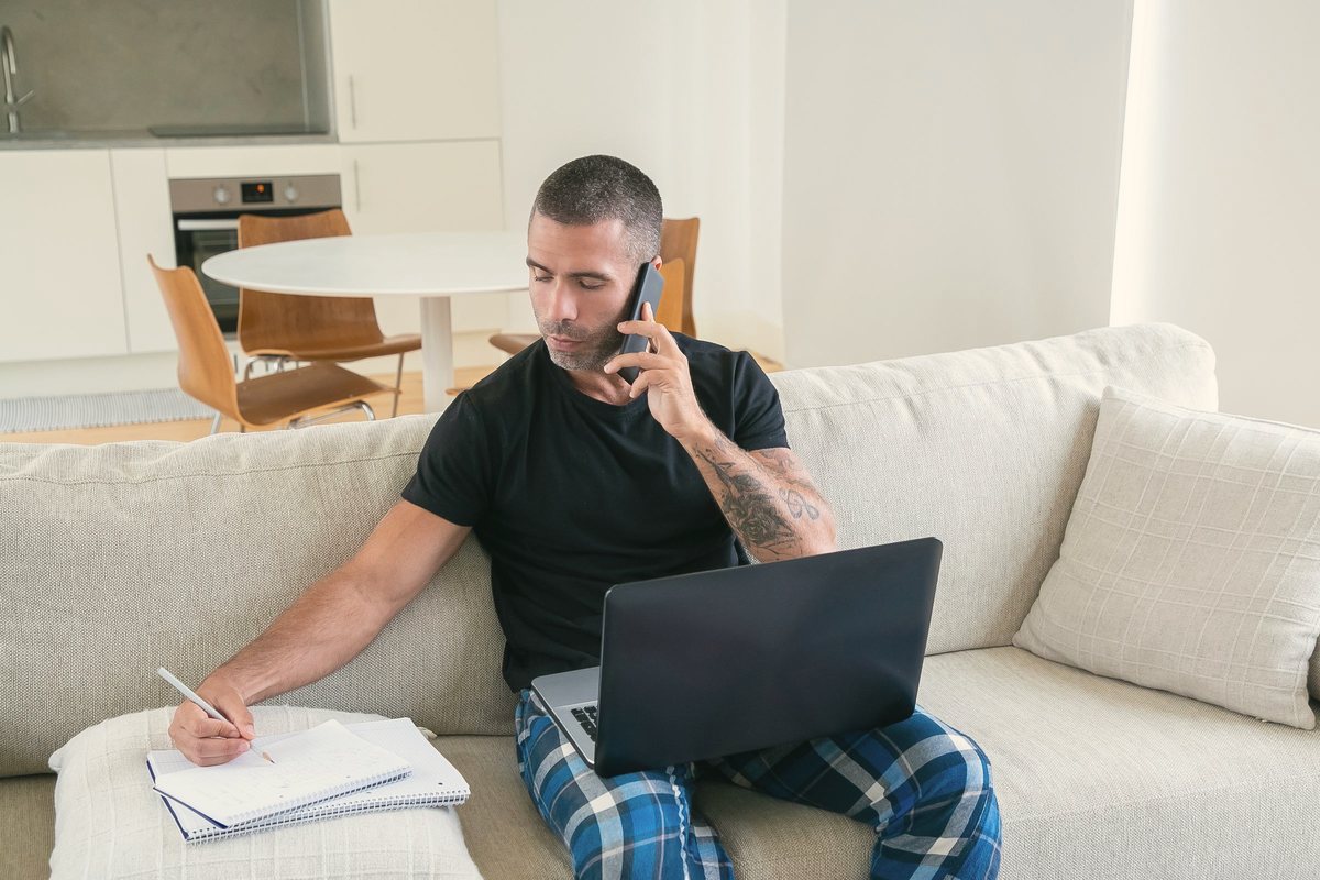 Man on a phone call at home taking notes on a notepad beside his laptop