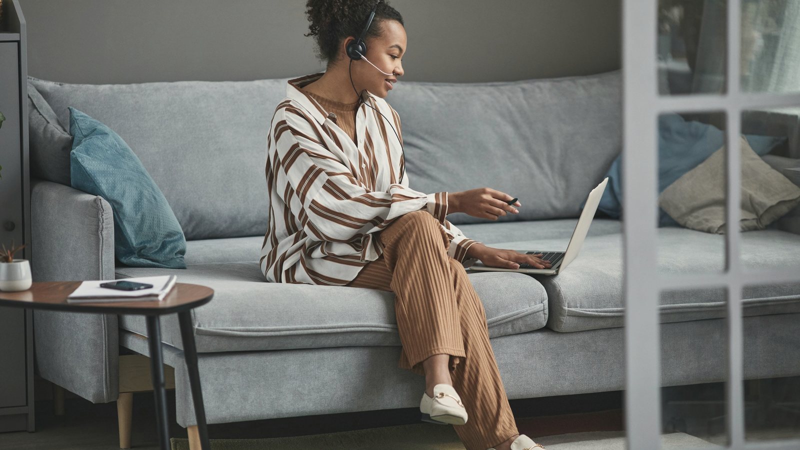 Woman on a sofa with a headset and laptop, joining a telehealth video call from home