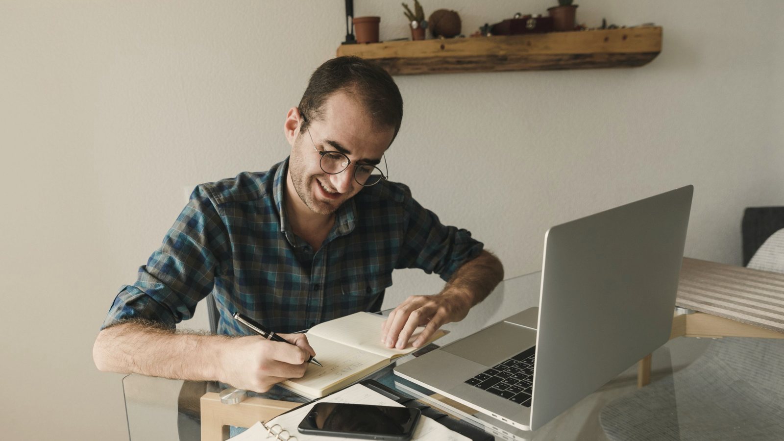 Man at a home desk writing in a notebook beside a laptop and phone
