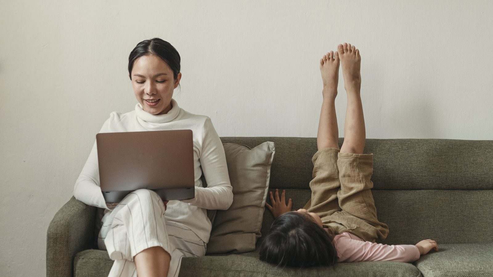 Cheerful mother working on laptop near daughter on couch