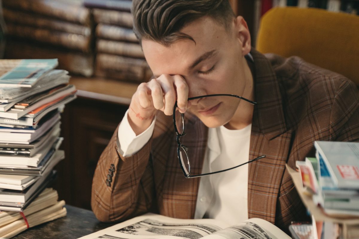 Tired student rubbing his eyes in front of a laptop surrounded by piles of books