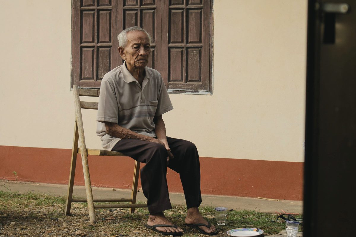 Older man sitting on a wooden chair outside a house