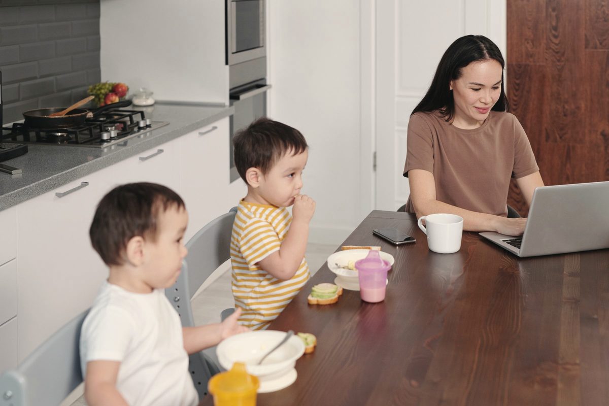 Woman working on her laptop while two kids eat breakfast beside her