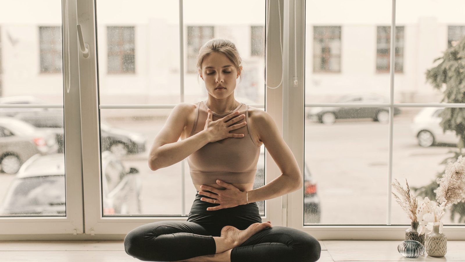A woman in leggings sitting near the glass windows while meditating