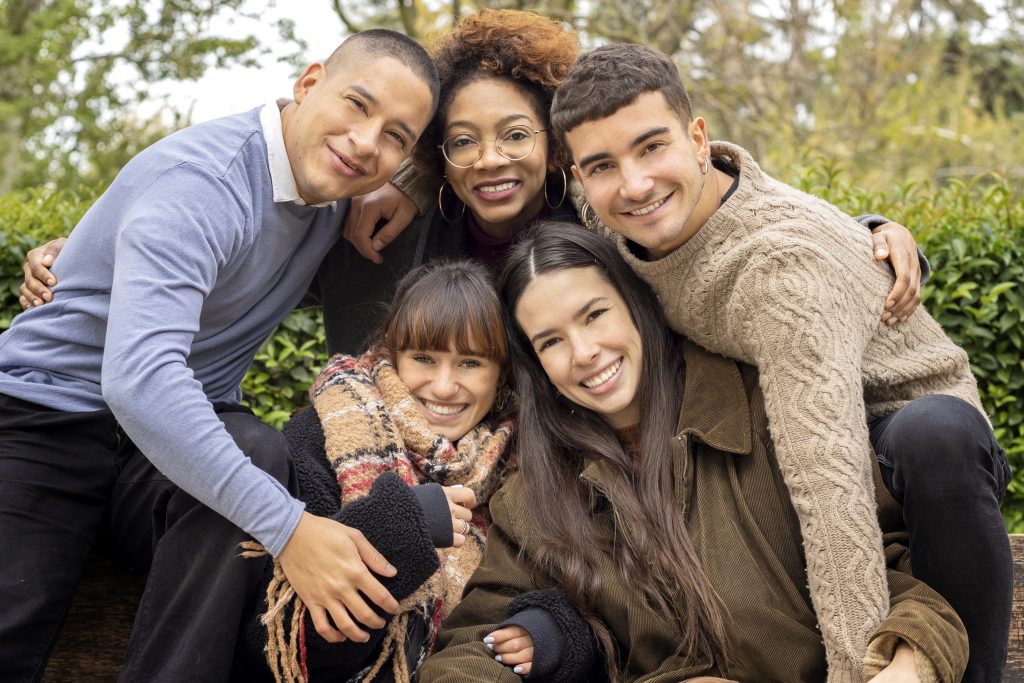 Five people sitting closely together outdoors, smiling at the camera in this sample post. Dressed in casual, warm clothing, they are surrounded by lush greenery.