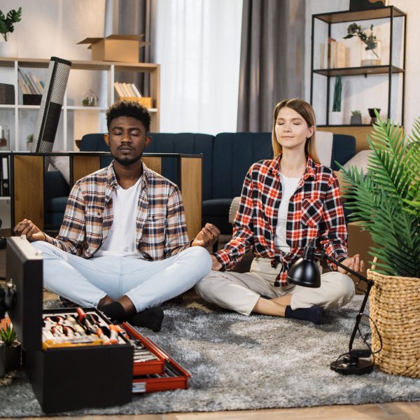 Two people sit cross-legged on a carpet, meditating amid the chaos of unpacked boxes and scattered tools, finding their guide to balanced living in the heart of their bustling living room.