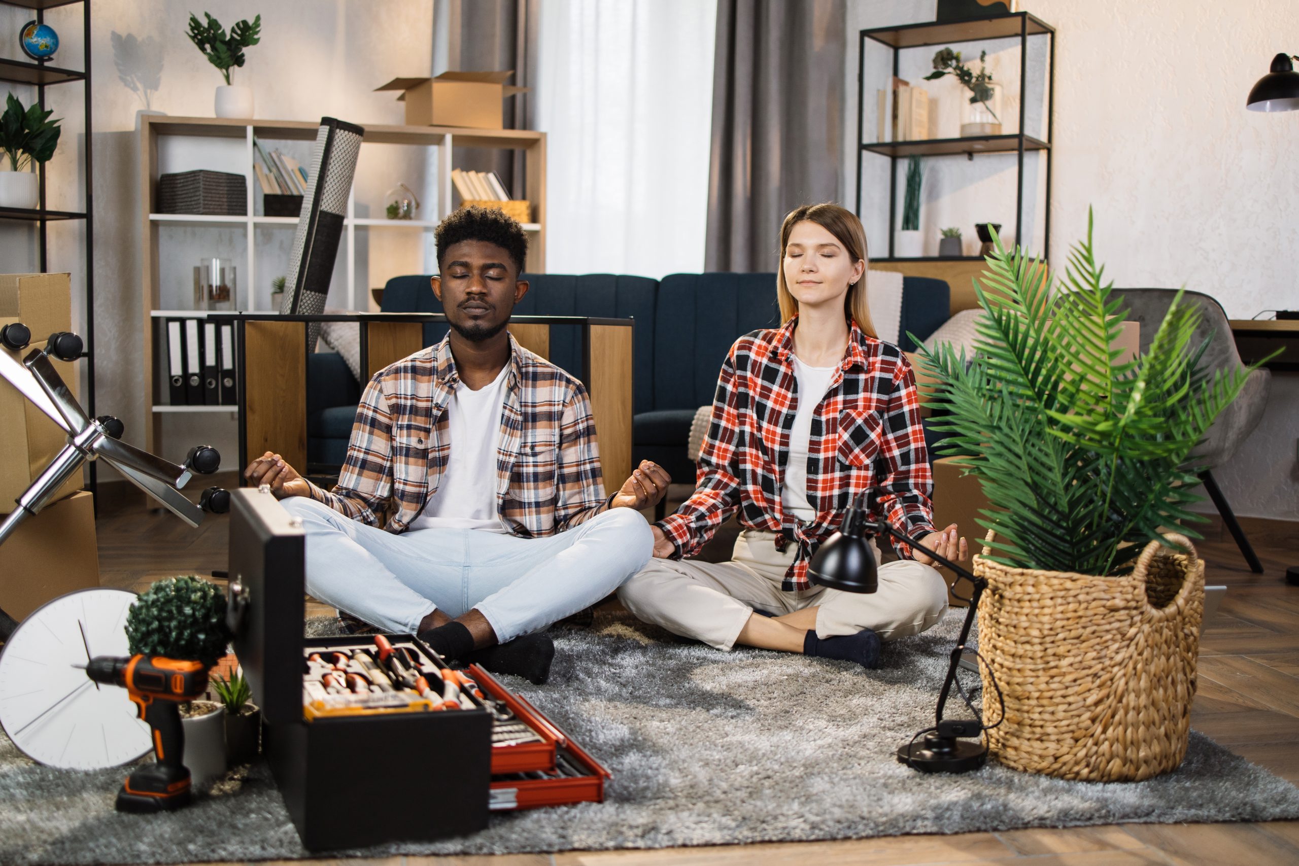 Two people sit cross-legged on a carpet, meditating amid the chaos of unpacked boxes and scattered tools, finding their guide to balanced living in the heart of their bustling living room.