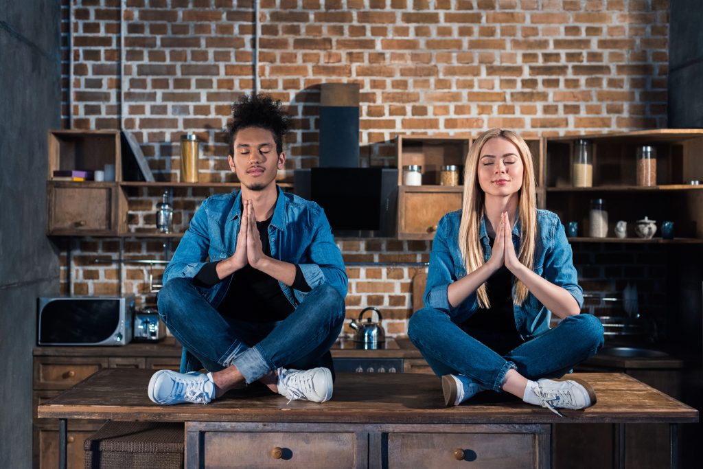 Two people sit cross-legged on a table in a brick-walled room, meditating with eyes closed and hands in prayer position. Both wear denim outfits and sneakers—a perfect Sample Post capturing tranquility amid urban chic.