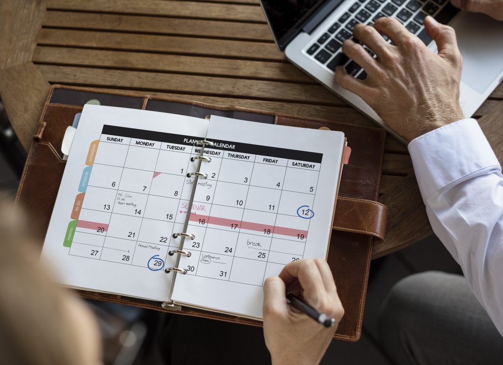 Person reviewing a physical planner with a circled date, alongside another person typing on a laptop—an ideal setup for Sample Post #2. The planner lies open on a wooden table, inviting curiosity and productivity in this cozy workspace scene.