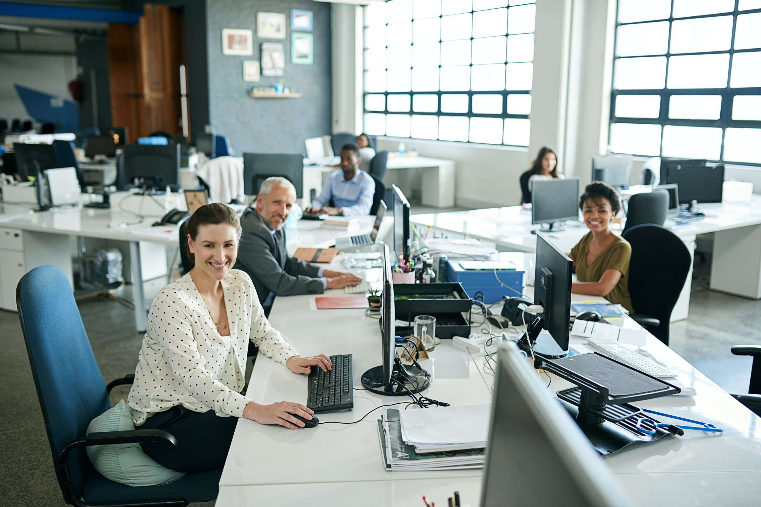 Office scene with five people efficiently working at desks adorned with computers, as large windows in the background flood the area with natural light, creating a vibrant atmosphere.