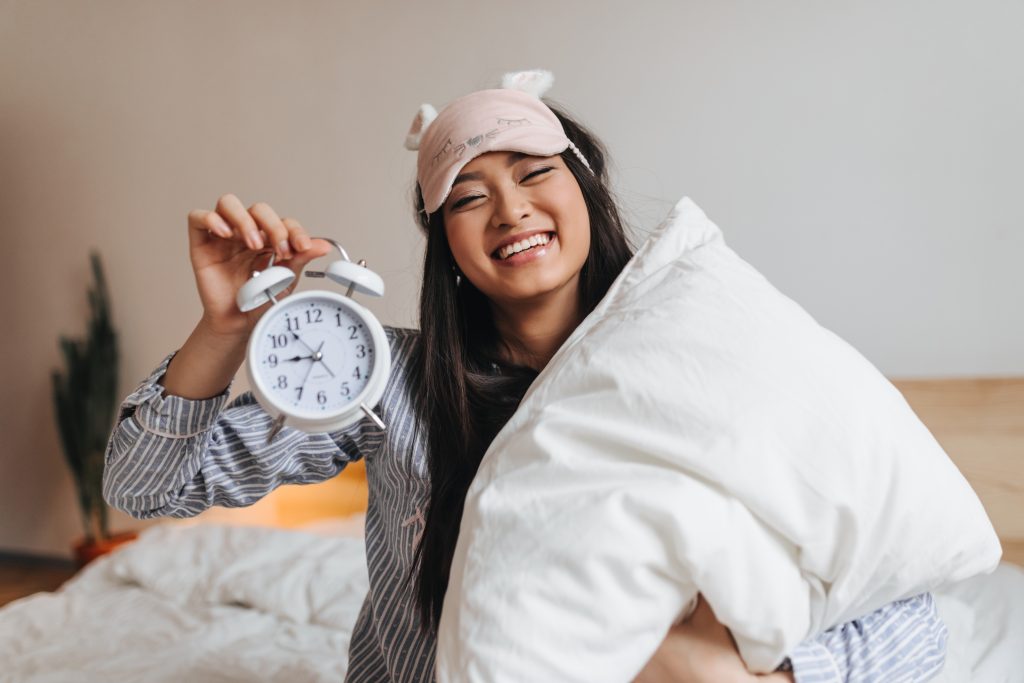 A person wearing pajamas and a sleep mask smiles while holding a pillow and an alarm clock, ready for the perfect Sample Post moment.