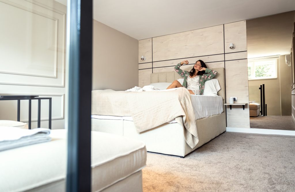 A woman sits on a bed in a modern, neutral-toned bedroom, stretching her arms. The room includes a mirror, a side table, and a window letting in natural light—a perfect scene for Sample Post #1.